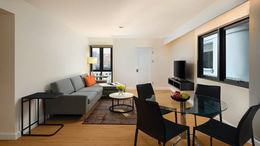 Modern living and dining area with a gray sofa, orange and white pillows, round glass dining table with black chairs, TV on the wall, large windows, wood flooring, and a small black side table.
