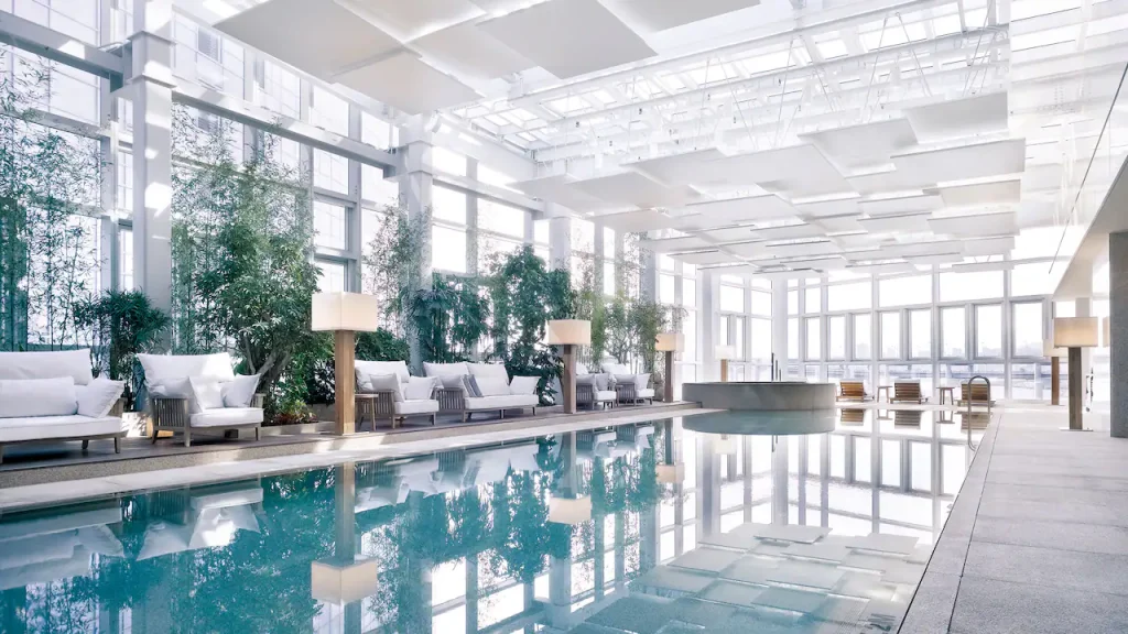 A bright, modern indoor pool area with large windows, white ceilings, lush green plants, and comfortable lounge seating along the sides. Natural light fills the space, creating a calm and inviting atmosphere.