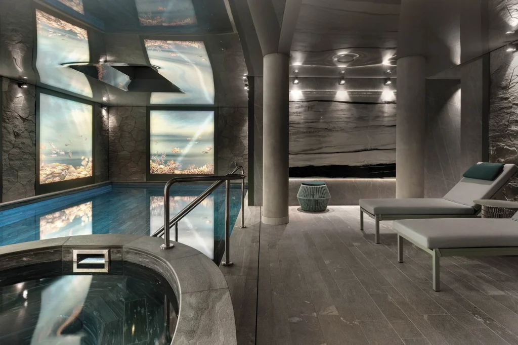 Luxurious indoor spa with a small pool, round hot tub, modern loungers, marble walls, columns, soft lighting, and large illuminated nature-themed wall panels.
