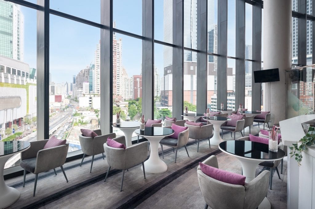 Modern indoor lounge with floor-to-ceiling windows, cityscape view, round tables, and cushioned chairs in neutral tones with purple pillows. Bright daylight fills the space, creating an open, inviting atmosphere.