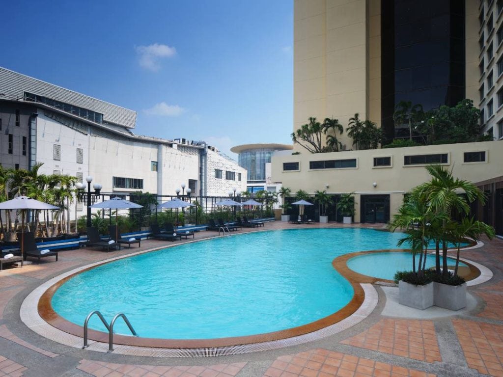 A circular outdoor swimming pool with lounge chairs and umbrellas, surrounded by palm trees, is set beside a tall building and other modern structures under a clear blue sky.