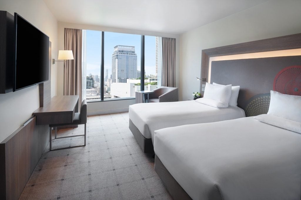 Modern hotel room with two double beds, a large wall-mounted TV, a desk with a chair, and a window offering a city view with tall buildings. The room features neutral colors and contemporary furnishings.