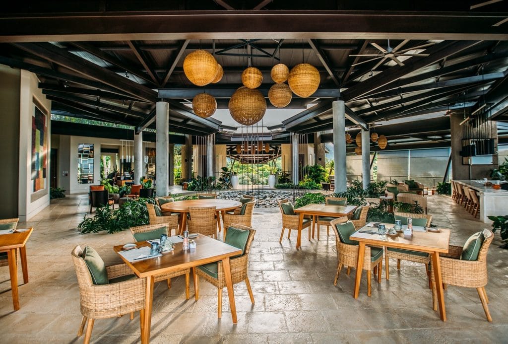 Open-air restaurant with wooden tables and wicker chairs set for dining. Large woven pendant lights hang from the ceiling, and lush greenery is visible inside and outside, creating a tropical, relaxed atmosphere.