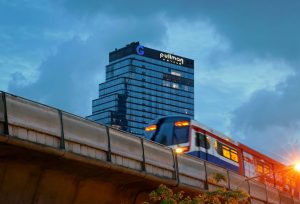 A blurred train moves along an elevated track at dusk, with the tall, modern Pullman Hotel building in the background against a cloudy sky.