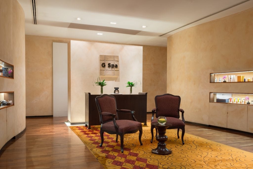 A spa reception area with two upholstered chairs and a small table on a yellow patterned rug, dark wood floors, shelves with products, and a sign reading G Spa on the back wall.