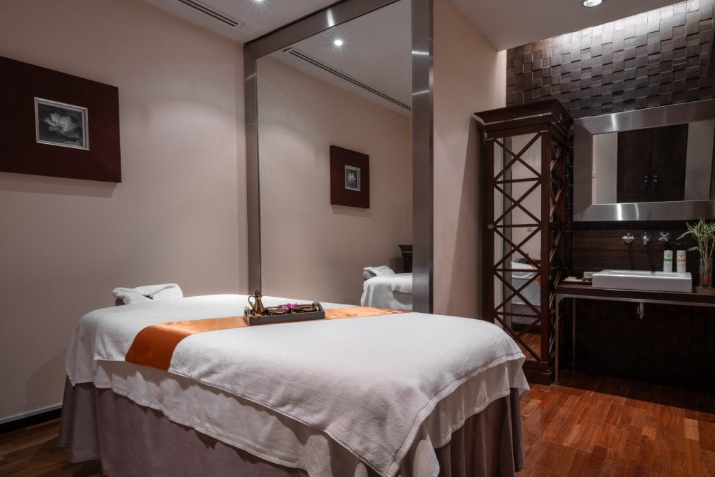 A tranquil spa room with a massage bed covered in white linens and an orange runner, a mirror, wall art, a dark wooden cabinet, a sink, and soft, warm lighting.