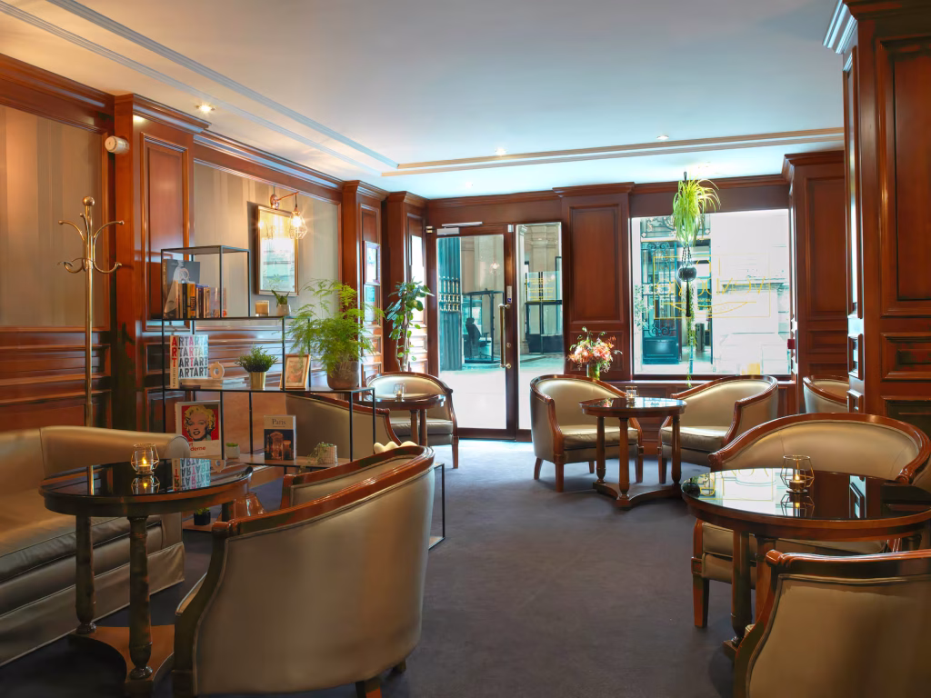 Elegant lounge with wood-paneled walls, glass tables, plush chairs, indoor plants, and bookshelves. Large windows and glass doors let in natural light, creating a cozy and inviting atmosphere.