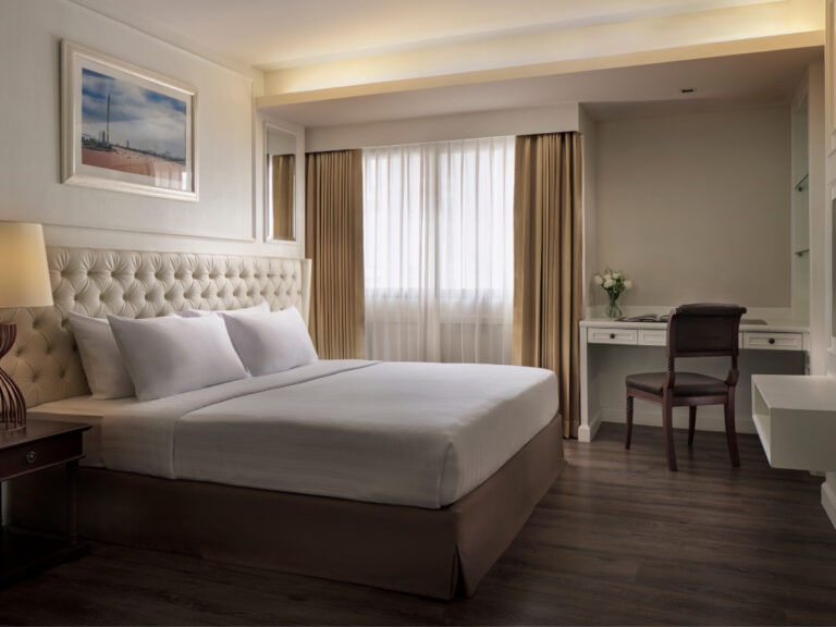 A modern hotel room with a large bed, white linens, a tufted headboard, a bedside lamp, a desk with a chair, and a vase of white flowers by the window with beige curtains. The floor is dark wood.