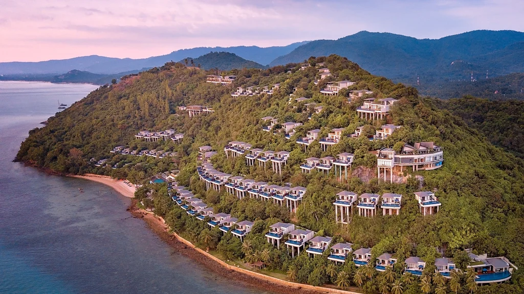 Aerial view of a lush, green hillside with modern villas and bungalows overlooking a coastline and sandy beach, with mountains in the background under a pastel-colored sky.