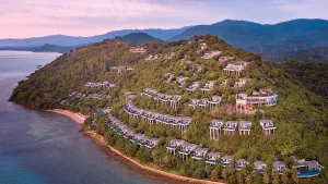 Aerial view of a lush, green hillside with modern villas and bungalows overlooking a coastline and sandy beach, with mountains in the background under a pastel-colored sky.