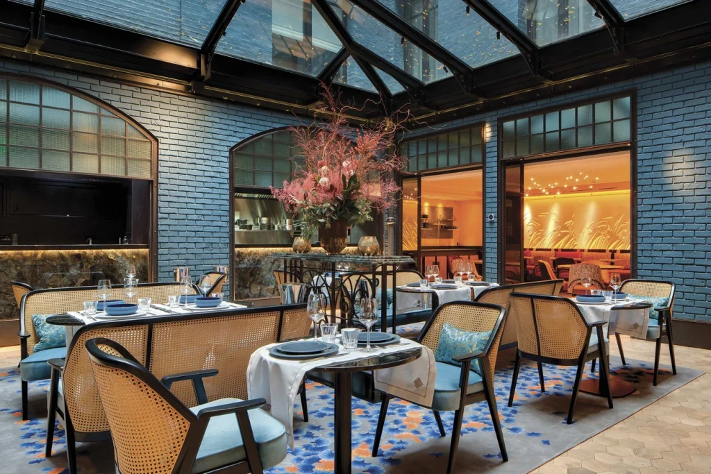 A stylish indoor dining area with blue brick walls, a glass ceiling, and elegant tables set with dishes and glasses. Wicker chairs surround the tables, and a large floral arrangement decorates the center. Warm lighting enhances the cozy ambiance.
