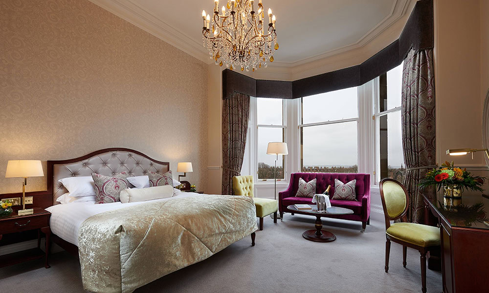 Elegant hotel room with a large bed, tufted headboard, and purple accents. A chandelier hangs from the ceiling, and there is a seating area by bay windows with a sofa, armchair, and a small table.