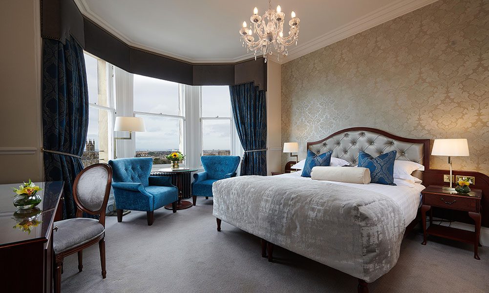Elegant hotel room with a large bed, blue chairs by tall windows, a chandelier, and ornate wallpaper. A desk with a chair and classic wooden furniture complete the sophisticated decor.