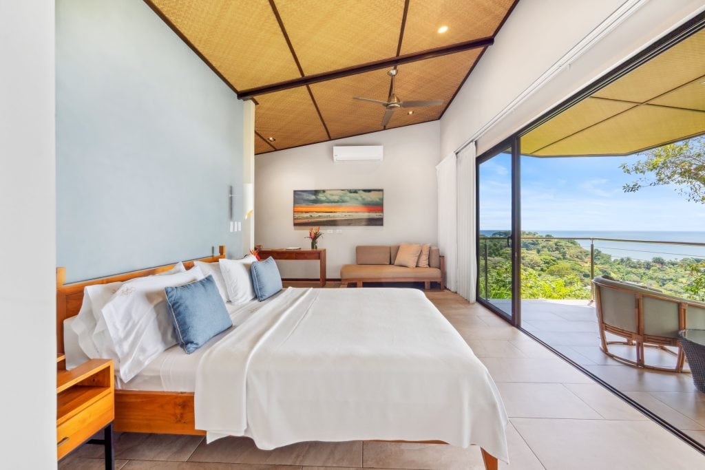 A modern, sunlit bedroom with a large bed, light blue accent wall, sofa, and glass doors leading to a balcony overlooking lush greenery and the ocean. A ceiling fan and wall art add to the airy, relaxing atmosphere.