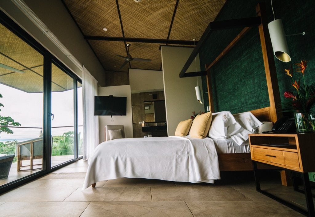 A modern hotel room with a large bed, white linens, decorative pillows, bedside tables, a wall-mounted TV, glass doors leading to a balcony, and a green accent wall. Natural light fills the space.