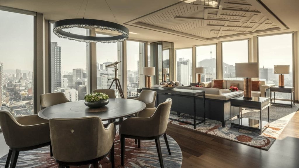 A modern, luxurious apartment with floor-to-ceiling windows offering city views, a round dining table with chairs, a stylish chandelier, a telescope, and a spacious living area with sofas and lamps.