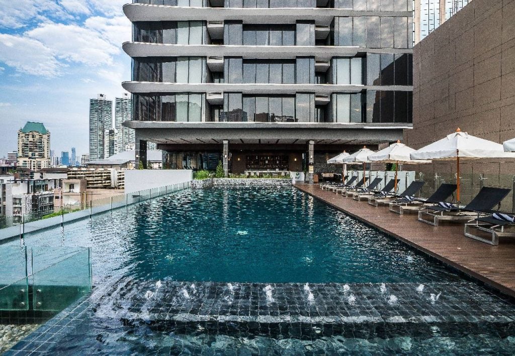 A modern rooftop swimming pool with lounge chairs and white umbrellas on a wooden deck, surrounded by tall buildings in an urban setting. The pool features a glass wall and a small waterfall at the front.