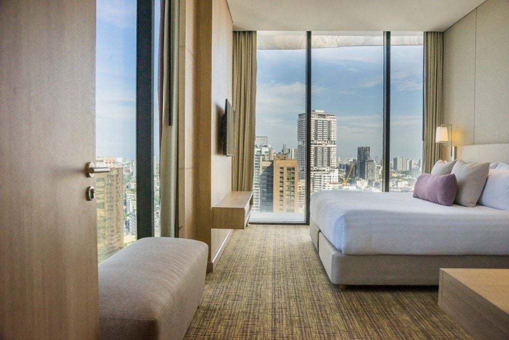 A modern hotel room with a large bed, light wood furniture, floor-to-ceiling windows, and a city skyline view with tall buildings under a blue sky.