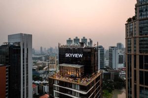 A rooftop bar named SKYVIEW sits atop a high-rise building, surrounded by other tall buildings in a cityscape at sunset, with people gathered on the rooftop terrace.