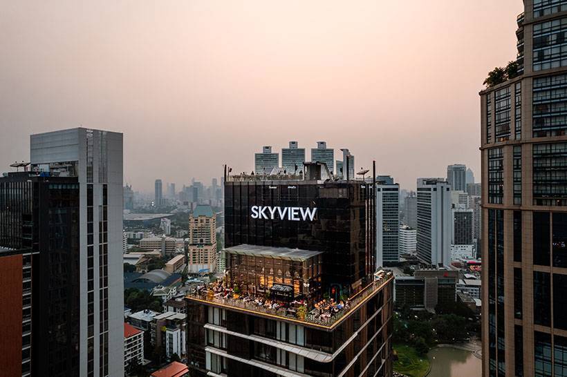A rooftop bar named SKYVIEW sits atop a high-rise building, surrounded by other tall buildings in a cityscape at sunset, with people gathered on the rooftop terrace.
