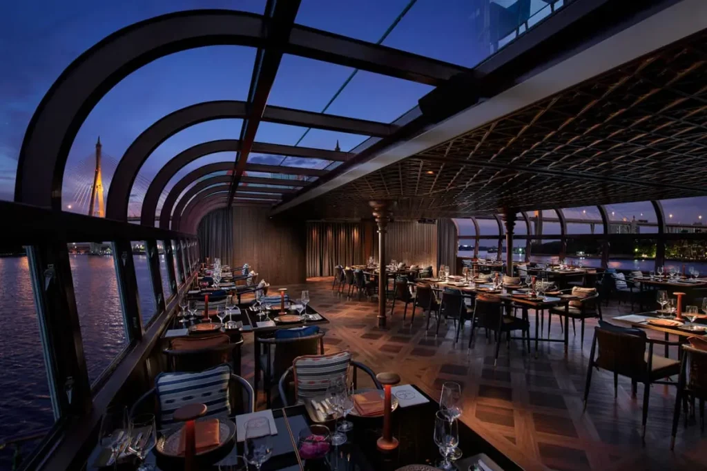 A stylish restaurant with floor-to-ceiling windows and a glass ceiling overlooks a river at sunset. Tables are set for dinner, and city lights and a bridge are visible through the windows. The ambiance is elegant and inviting.