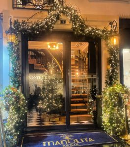 A warmly lit entrance to Manolita Brunch, decorated with festive garlands and string lights. The glass doors reveal a spiral staircase and a Christmas tree inside. A blue doormat with the Manolita logo lies at the door.