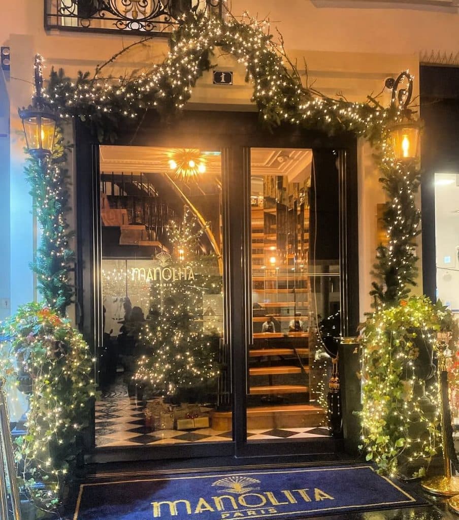 A warmly lit entrance to Manolita Brunch, decorated with festive garlands and string lights. The glass doors reveal a spiral staircase and a Christmas tree inside. A blue doormat with the Manolita logo lies at the door.