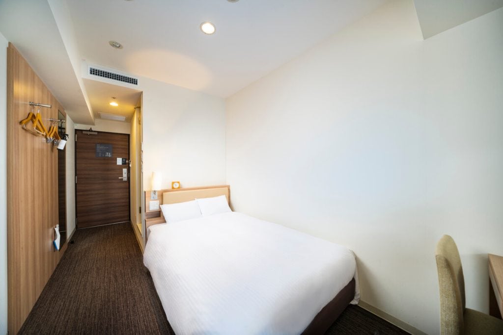 A compact hotel room with a double bed, white bedding, a wooden headboard, wall-mounted bedside light, small desk with a beige chair, and an entryway with a dark wooden door and coat hangers on the wall.