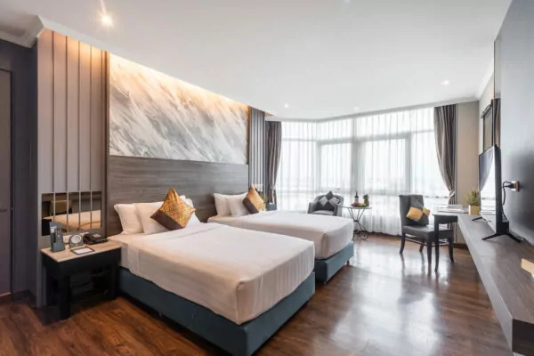 A modern hotel room with two neatly made double beds, a marble-patterned accent wall, large windows with sheer curtains, a desk with a chair, wooden floors, and contemporary decor.