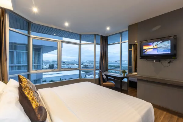 Modern hotel room with large windows offering a scenic city and water view, featuring a neatly made double bed, wall-mounted TV, desk, chair, and soft lighting.