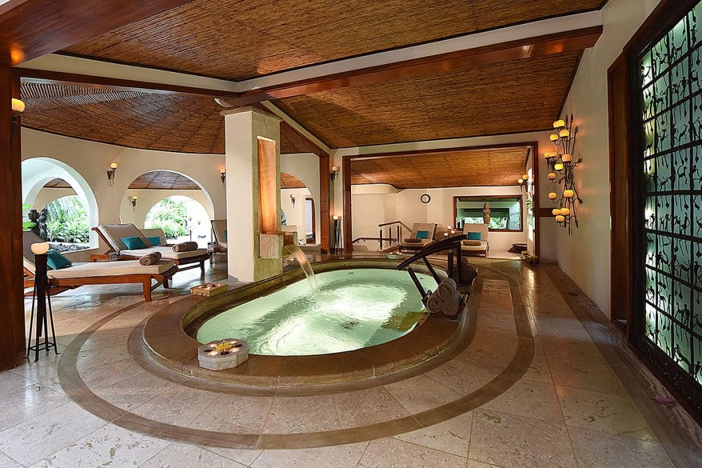 A luxurious indoor spa with a round soaking pool, wooden beams, lounge chairs, candles on the walls, and large windows revealing lush greenery outside. The space features soft lighting and a relaxing ambiance.