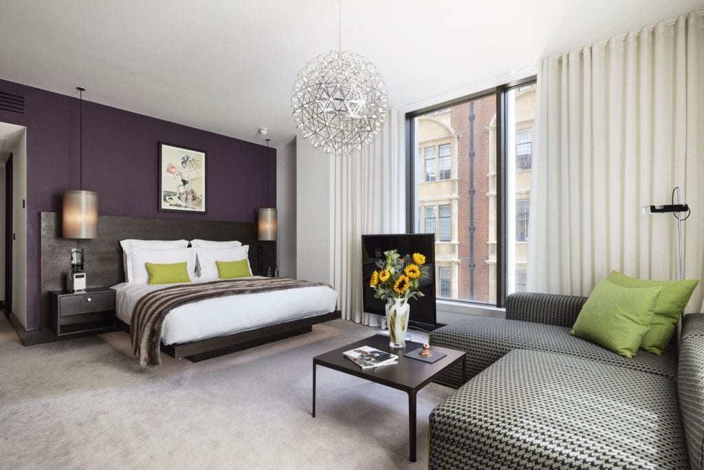 Modern hotel room with a large bed, purple accent wall, two green pillows, a couch with green cushions, a coffee table with a vase of sunflowers, and large windows with white curtains.