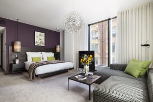 Modern hotel room with a large bed, purple accent wall, two green pillows, a couch with green cushions, a coffee table with a vase of sunflowers, and large windows with white curtains.
