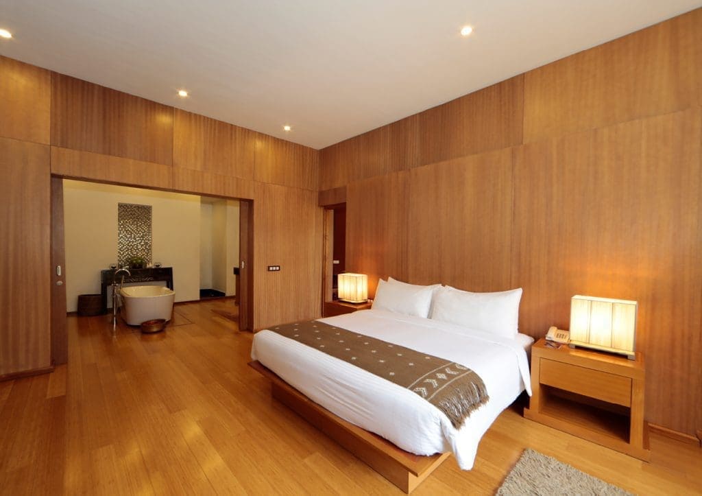 Spacious hotel room with wooden walls and floor, a large bed with white linens, two bedside tables with lamps, and an open doorway leading to a bathroom with a bathtub.