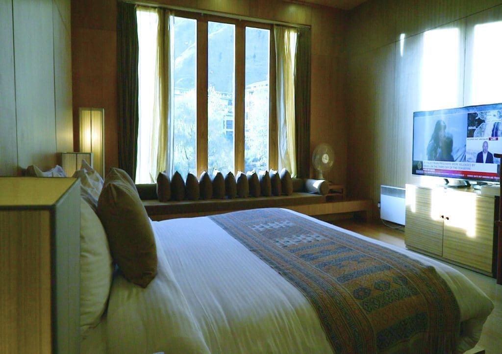 A cozy hotel room with a large bed, decorative pillows, a wooden bench by tall windows with curtains, a flat-screen TV on a cabinet, and sunlight streaming in.
