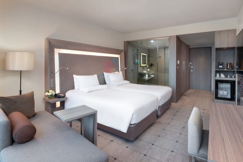 Modern hotel room with two twin beds, a glass-walled bathroom, a sofa, desk, lamp, minibar, and coffee station. Neutral tones and soft lighting create a clean, comfortable atmosphere.