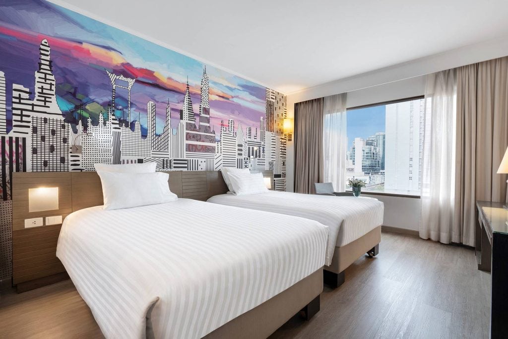 Modern hotel room with two single beds, striped white bedding, a large colorful cityscape mural on the wall, and a window with city views letting in natural light.