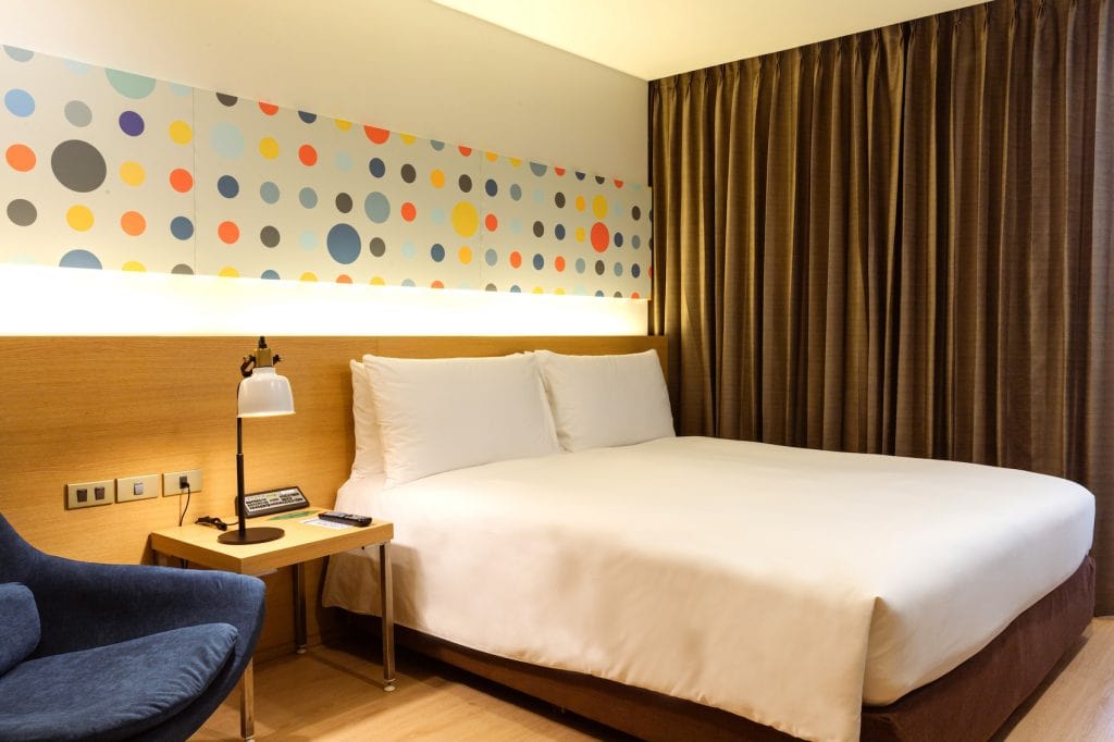 A modern hotel room with a large bed, white bedding, a wooden headboard, a side table with a lamp and phone, a blue chair, brown curtains, and a colorful polka dot wall decoration above the bed.