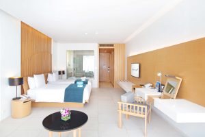 Modern hotel room with two double beds, teal bed runners, a round black table with a flower vase, a wooden accent wall, desk area, and a glass-walled bathroom in the background. Room is bright and minimalistic.