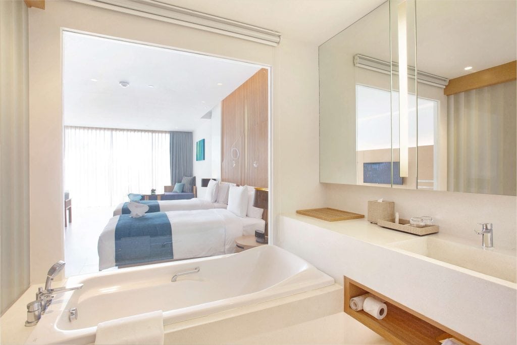 A modern hotel room with two beds, blue accents, a large window with sheer curtains, and a view from the bathroom featuring a bathtub, sink, and mirror. The design is bright and minimalist.