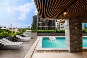Outdoor pool with a rainbow-striped inflatable float, surrounded by lounge chairs. Modern buildings and greenery are visible in the background under a partly cloudy sky.