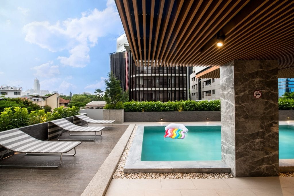 Outdoor pool with a rainbow-striped inflatable float, surrounded by lounge chairs. Modern buildings and greenery are visible in the background under a partly cloudy sky.