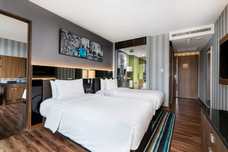 Modern hotel room with two white twin beds, striped area rug, wood floors, a large mirror, wall art above the beds, and a view of the entryway and bathroom in the background.