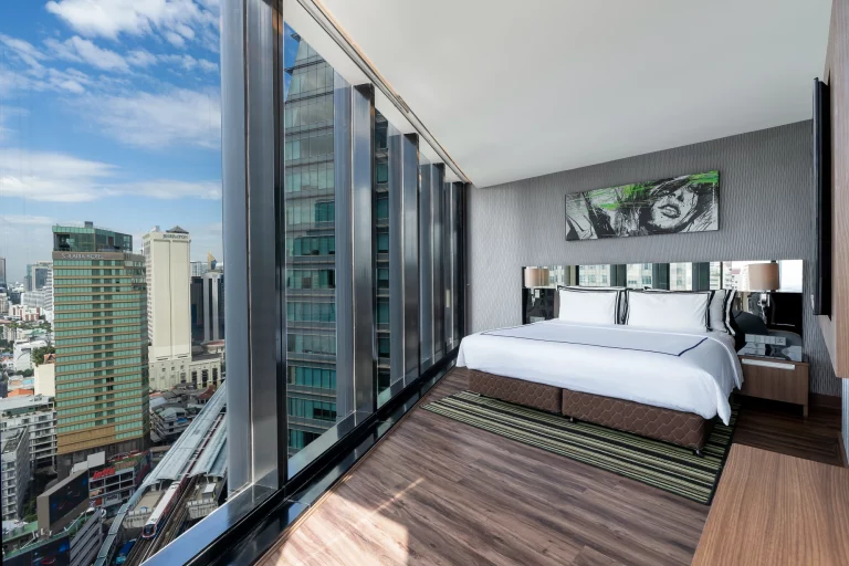 Modern hotel room with a large bed and floor-to-ceiling windows offering a city skyline view. The room has wood flooring, minimal furnishings, and contemporary decor with a green and black artwork above the bed.