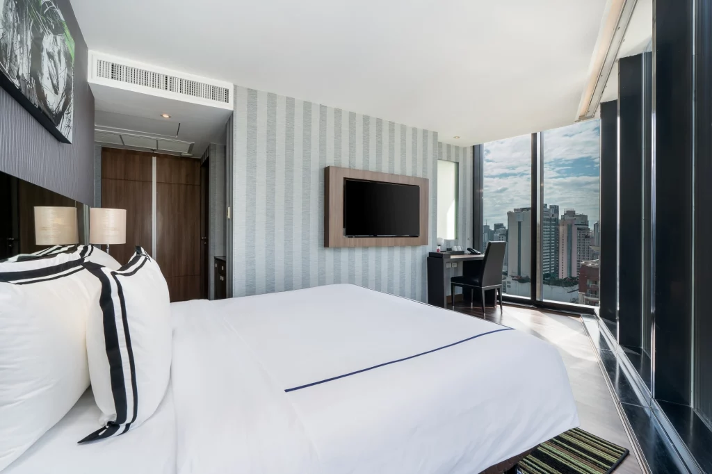 A modern hotel room with a large bed, striped pillows, wall-mounted TV, desk and chair by floor-to-ceiling windows, and a city skyline view outside. Light decor with patterned wallpaper and wooden accents.