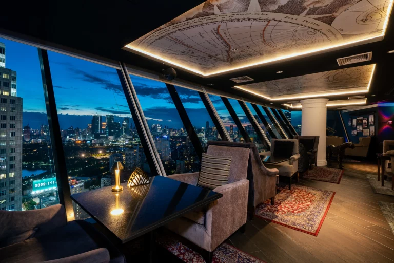 A modern rooftop lounge with plush chairs, tables, and large windows offering a panoramic view of a city skyline at dusk, with warm ambient lighting and a decorative map on the ceiling.