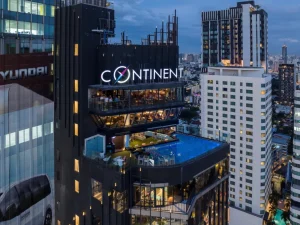 A modern high-rise building with CONTINENT signage features a rooftop infinity pool and outdoor lounge area, overlooking a cityscape with numerous tall buildings at dusk.