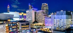 Cityscape at night featuring modern high-rise buildings with illuminated windows, including a prominent Novotel hotel in the center and other towers in the background against a deep blue sky.