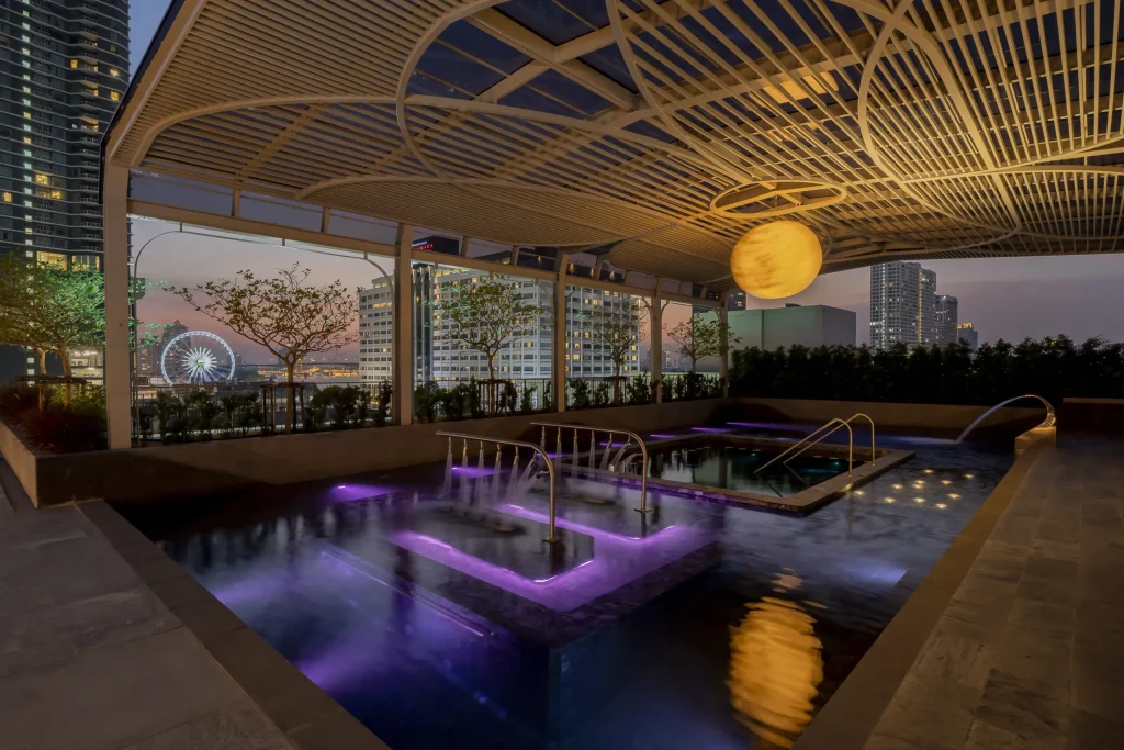 A modern rooftop pool area at dusk, featuring glowing purple-lit pools, a large circular light fixture overhead, and city skyscrapers in the background with a ferris wheel visible.
