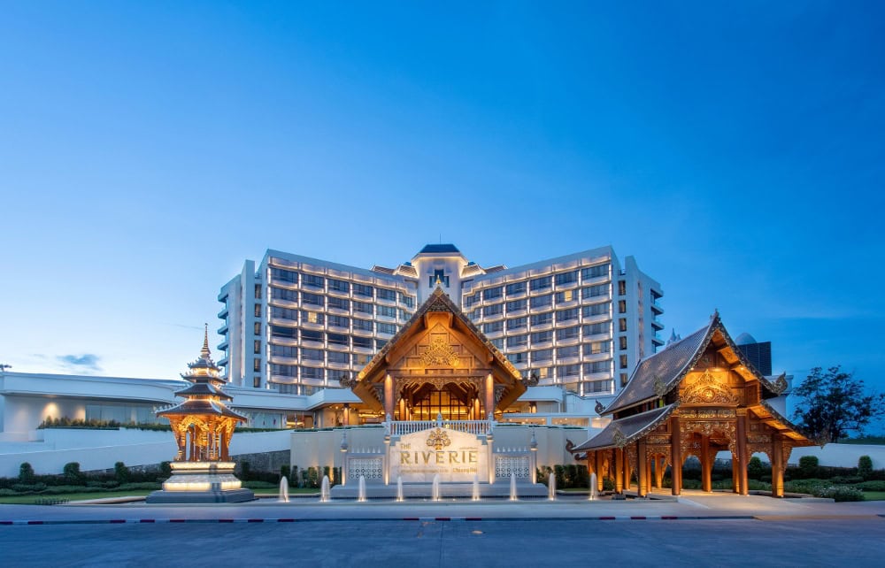 A large, modern hotel with balconies stands behind ornate, traditional Thai pavilions and a sign reading The Riverie during twilight, under a clear blue sky.